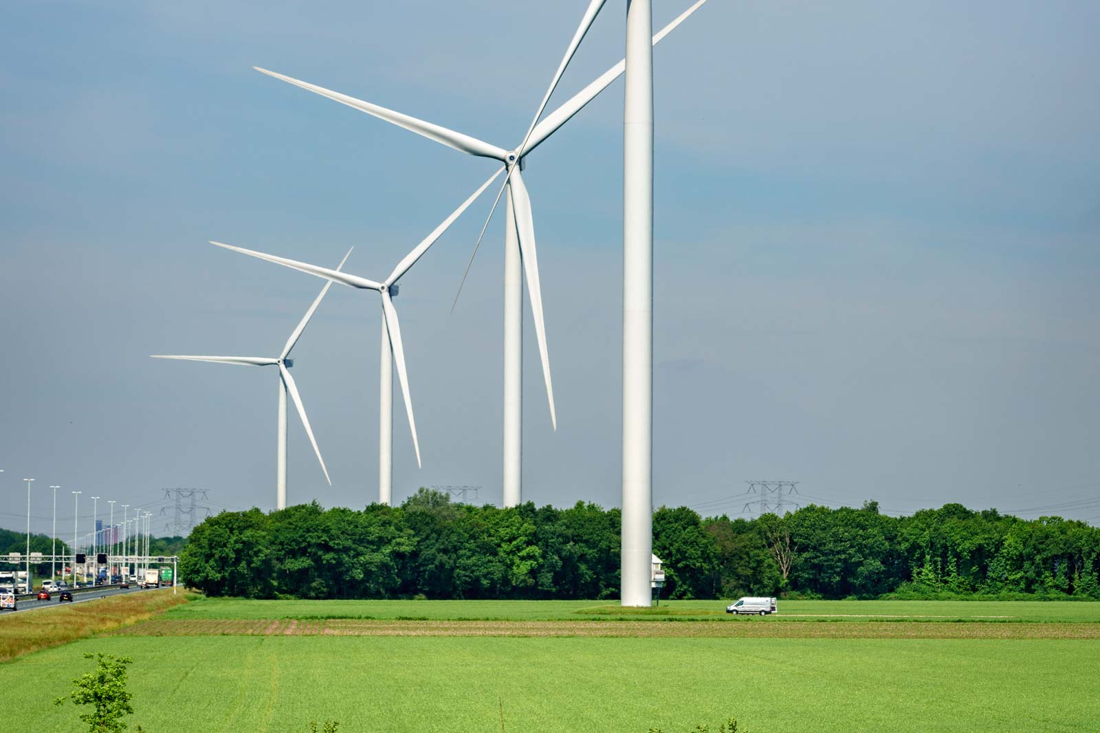 Een landschap met hoge windturbines naast een snelweg en groene velden onder een heldere blauwe lucht.