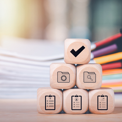Een stapel houten blokken met iconen die goedkeuring, instellingen, zoeken en checklists voorstellen, op een bureau met stapels papier.