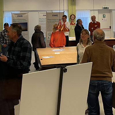 Een groep mensen die in gesprek is op een evenement, met een grote tafel en informatieve banners op de achtergrond.